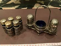 Photo showing group of four antique English condiment shakers with brass tops and blue glass liners beside twin shaker set in metal stand with handle.