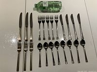 Two partial vintage cutlery sets laid out on a white surface with a green water bottle for size reference. Each set includes 6 knives, forks, and spoons neatly arranged in rows.