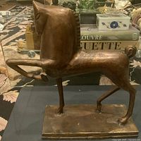 Side profile of the stylized metal Trojan Horse sculpture showing the horse's head, curved neck, and front leg extended forward, mounted on a rectangular base.