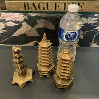 Three vintage brass pagoda decor items shown with water bottle for scale. Two items are a matched pair with one missing the tip. The third pagoda also missing its tip. Hexagonal bases and tiered roofs are visible.
