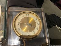 Front-facing photo of the vintage Seth Thomas lucite clock showing the square transparent lucite body and round gold-tone clock face with black Arabic numerals and gold hands.