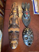 Three masks displayed on wooden table showing tribal and geometric patterns including bead designs