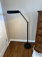 Full view of the black adjustable standing floor lamp with cylindrical lampshade and round base on wooden floor near wall and door.