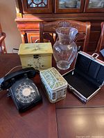 Black rotary phone, decorative metal tin, wooden recipe box with floral design, clear glass vase, and pink case with buckle on table