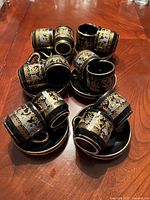 Twelve espresso cups and saucers displayed on a wooden table showing black ceramic with intricate 24k gold floral and ornamental designs.