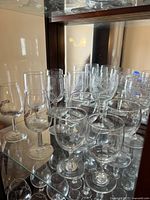 Photo showing 12 assorted clear glass wine glasses displayed on shelves. Glasses vary in height and bowl shapes, some with rounded bowls and others taller and narrower. Minor surface wear visible but no cracks or chips.