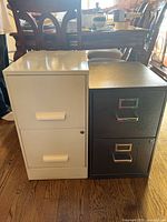 Photo shows two small filing cabinets side-by-side on wooden floor in an indoor setting with other furniture in the background.