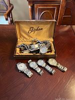 Box with 'Brigham' branding on lid, containing multiple watches with metal bands, and 5 watches arranged outside the box on a wooden table.