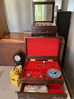 Photo showing two large wooden jewelry boxes with compartments and red interior lining, open to reveal small jewelry items, along with yellow lion bookends, black clock, blue collector's tin, and an illuminated magnifying glass on a flat surface.