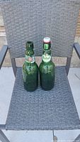 Two green Grolsch glass swivel top bottles placed on outdoor chair seat. One bottle has cap attached and closed, the other bottle is open without top cap.