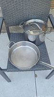 Front view of clean stainless steel pots and frying pan with lids on dark woven chair.