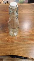 Clear glass bottle with white ceramic flip-top lid and metal clasp on wooden table.