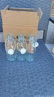 Photo showing three clear glass swing top bottles with plastic lids and rubber gaskets displayed in front of a cardboard box outdoors on a textured table surface.