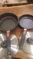 Photo showing two black fry pans side by side with removable handles and stainless steel lids below them.