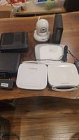 Photo of all devices on a wooden surface: includes one white IP dome security camera with antenna, two black modem/router units, three white wireless router/modem units, and one power supply.