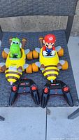 Two yellow bee-themed remote control cars with Mario and Yoshi figures, each with separate black remote controllers on a chair.