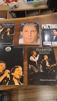 Five Paul Simon and Simon & Garfunkel related books arranged on a wooden surface, showing front covers including biographies and a photo portrait of Paul Simon.