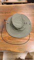 Top-down view of olive green Tilley sun hat with wide brim and button details on crown. Black and gray adjustable chin lanyard shown detached.