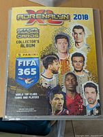 Yellow cover of the Adrenalyn XL 2018 official FIFA 365 collector's album showing images of six soccer players on front cover.
