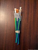 Bundle of small painting brushes with various sized flat, angled, and round bristles on wooden handles painted green, blue, and yellow, held with a rubber band on dark wooden surface.