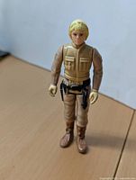 Front view of Luke Skywalker action figure showing tan flight suit, blonde hair, belt with black holster, and boots.