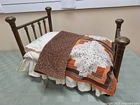 Antique brass doll's bed made of brass with four posts and rounded finials, dressed with quilt, pillow, and sheets.