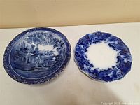 Both flow blue ceramic plates displayed together on a white surface, showing difference in designs and scalloped edges.