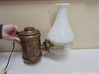 Full view of the antique electric lamp showing embossed metal body, glass reservoir, and white glass chimney.