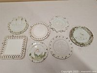 Seven antique milk glass plates arranged on flat surface showing variety of edge styles and embossed images including birds, chickens, and owls.
