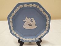 Octagonal blue jasperware plate with white classical relief figures in center and white leaf wreath border. Plate is displayed on black stand against neutral background.