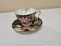 Side view of Aynsley bone china teacup and saucer showing floral design, black background, scalloped edges with gold trim, and lotus flower motif.