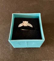 Fashion ring displayed in teal box, showing front view of sparkling clear crystal stone setting on silver-tone band.