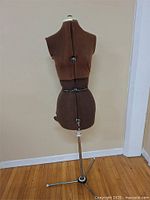 Full view of the brown dressmaker's form on metal tripod stand against a beige wall and hardwood floor.