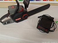 Electric Oregon 40V Lithium Ion chainsaw next to black battery charger with cords, resting on a white surface.