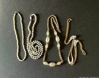 Four necklaces laid flat on black background, showing variety in length and style including white faux pearls, gold-tone chains, and multicolored pearls