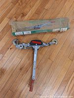 Cable puller device laid flat on wood floor with original box above it, showing handle, hooks, and central mechanism.