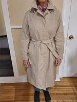 Full front view of beige women's trench coat with belt, worn by person, showing length down to knees, collar, and pockets.