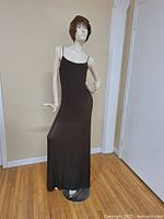 Full view of the mannequin standing against a beige wall on a hardwood floor, wearing brown ribbed dress and wig.