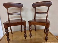 Two matching antique wooden chairs, side by side, showing full front and back view with turned legs, curved backrest, and flat seat.