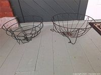 Two metal hanging plant holders placed on a porch with a gray door as background, showing size and condition.