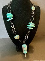 Full view of the necklace displayed on a black bust, showing turquoise dyed howlite nugget beads, black hammered and twisted metal links, and central abstract ceramic and stone pendant.