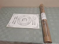 Paper placemat laid flat on a table next to unopened roll of paper table runner wrapped in brown label with cutlery illustrations.