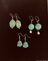 Top-down shot of all three pairs of earrings arranged on a dark background showing oval stone beads, round beads with pearls, and carved Buddha face beads.