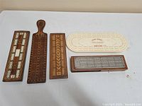 Four vintage cribbage boards laid out on a table: three wooden with detailed inlays including one with a handle and one plastic Royal Crown board. Also, a small wooden box with metal dominoes is present.