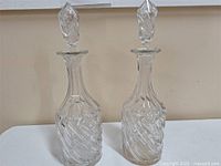 Two vintage cut-glass decanters with intricate swirling patterns and matching stoppers, shown side by side on white surface.