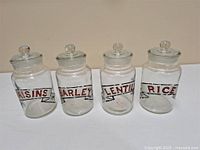 Front view of four vintage glass canisters labeled Raisins, Barley, Lentils, and Rice with clear glass lids with round knob handles, arranged in a row.