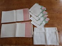 Lot of six hand towels with pink horizontal stripes and a stack of cream serviettes with embroidery on table.