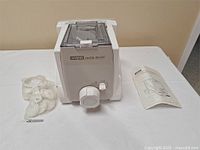 White Waring Pasta Maker unit with accessories in plastic bag and instruction manual next to it on table.