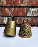 Two brass bells side by side on white surface against brick wall. Heavier thicker sanctuary bell on left with clip but no internal ringer visible. Ornate cow bell on right with raised animal detail and loop handle on top.