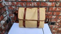 Front view of beige canvas messenger bag with brown leather trim and buckle straps against a brick background.